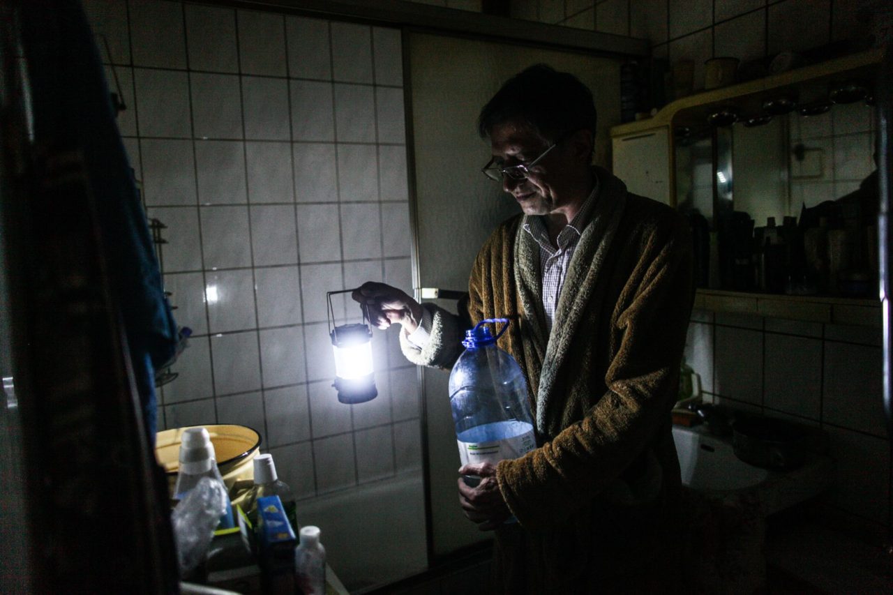 Владимир в ванной комнате занимается хозяйственными делами, воду и свет отключили, поэтому вся вода из пятилитровых бутылок, а вместо света маленький фонарик